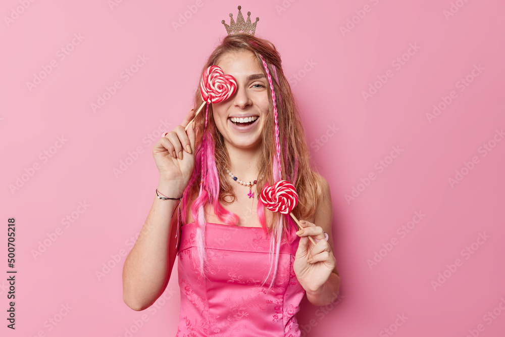 Positive European woman covers eye with heart shaped lollipop wears festive dress and small crown on head smiles broadly has white perfect teeth poses against pink background. Time for party