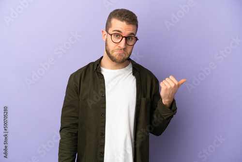 Young handsome caucasian man isolated on purple background unhappy and pointing to the side