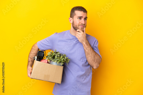 Young caucasian making a move while picking up a box full of things isolated on yellow background having doubts