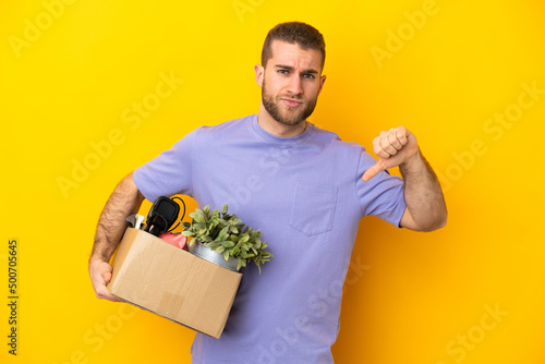 Young caucasian making a move while picking up a box full of things isolated on yellow background showing thumb down with negative expression