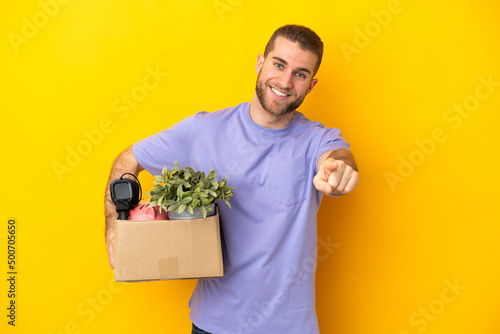 Young caucasian making a move while picking up a box full of things isolated on yellow background pointing front with happy expression