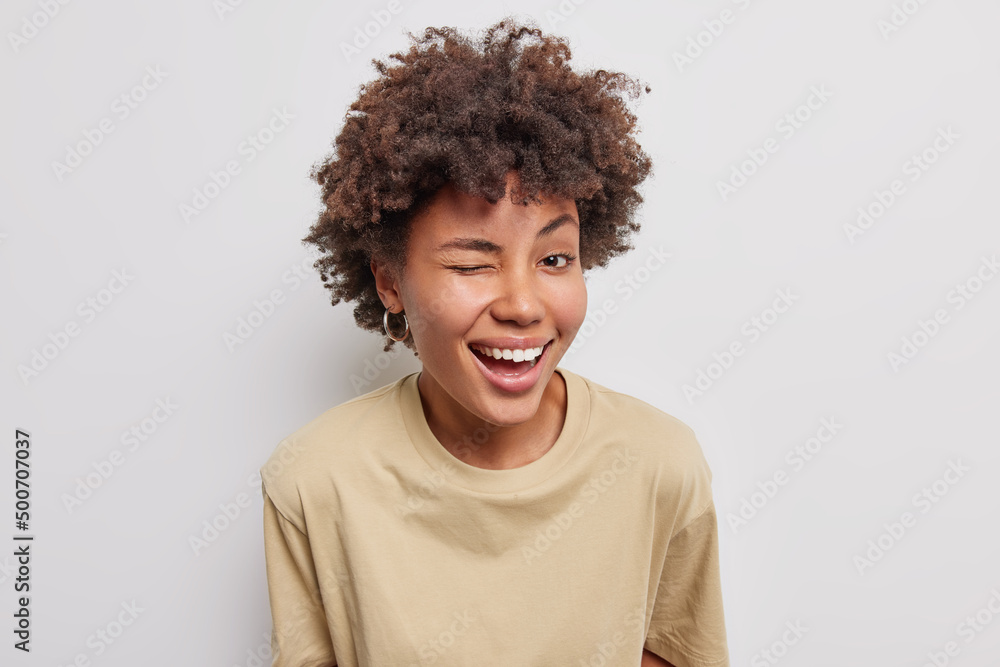 Friendly cheerful woman has playful mood winks eye smiles broadly dressed in casual beige t shirt dressed in casual brown t shirt isolated over white background. Positive human emotions concept Friendly cheerful woman has playful mood winks eye smiles broadly dressed in casual beige t shirt dressed in casual brown t shirt isolated over white background. Positive human emotions concept
