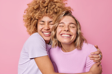 Cheerful young women embrace each other with love feel very glad smile broadly show white teeth care about each other dressed in casual t shirts isolated over pink background. Friendship concept