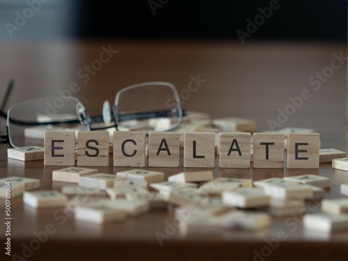 escalate word or concept represented by wooden letter tiles on a wooden table with glasses and a book photo