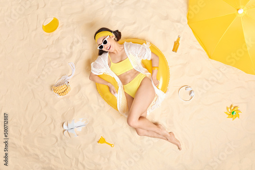 Young brunette woman has cheerful expression wears sunglasses and yellow swimsuit poses on inflated swimring spends free time at beach gets suntan during tropical vacation. Summer time concept