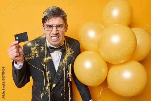 Irritated man clenches teeth looks angrily holds banking card and bunch of inflated balloons dressed in festive outfit feels outraged as hasne enough money for celebrtion. Party time concept photo