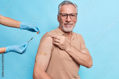 Vaccine injection against coronavirus. Pleased old man being immunized against covid 19 at hospital during visit to doctor isolated over blue wall wears plaster on arm. Vaccination for elderly people