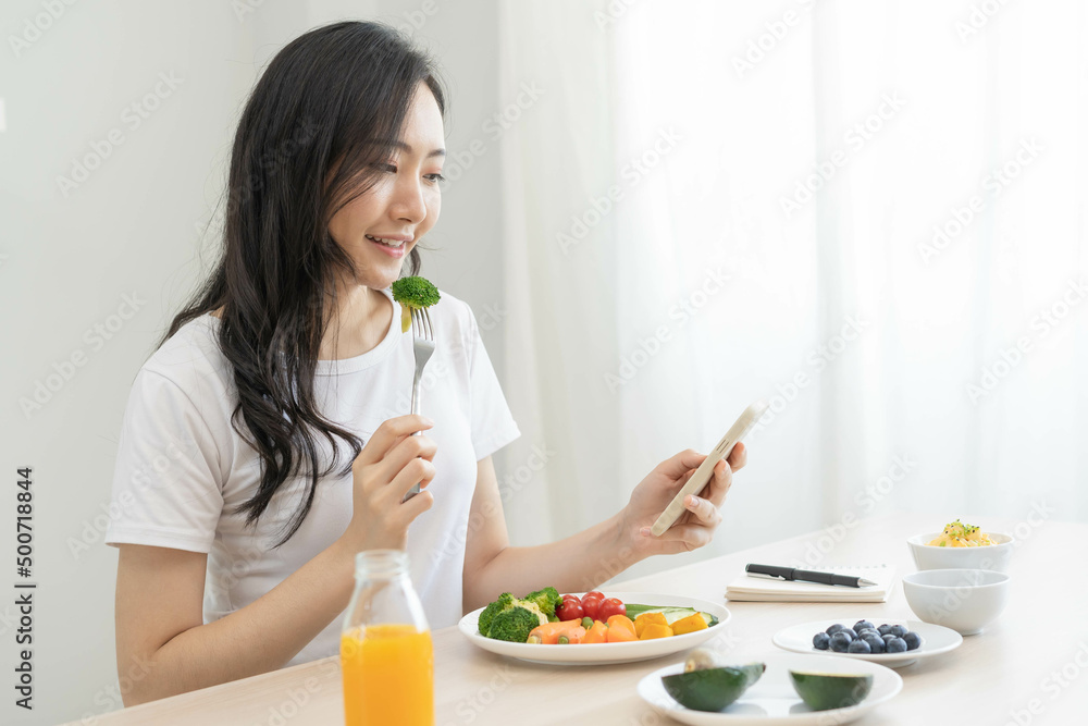 Dieting, asian young woman eating, holding fork at broccoli, diet plan nutrition with fresh vegetables salad, enjoy meal while using smartphone. Nutritionist of healthy, nutrition of weight loss. - obrazy, fototapety, plakaty 
