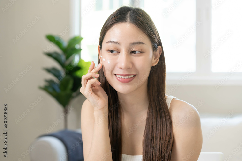 Fresh healthy skin, beautiful smile of asian young woman, girl looking at mirror, applying moisturizer on her face, putting cream treatment before makeup cosmetic routine at home. Facial Beauty. Fresh healthy skin, beautiful smile of asian young woman, girl looking at mirror, applying moisturizer on her face, putting cream treatment before makeup cosmetic routine at home. Facial Beauty.