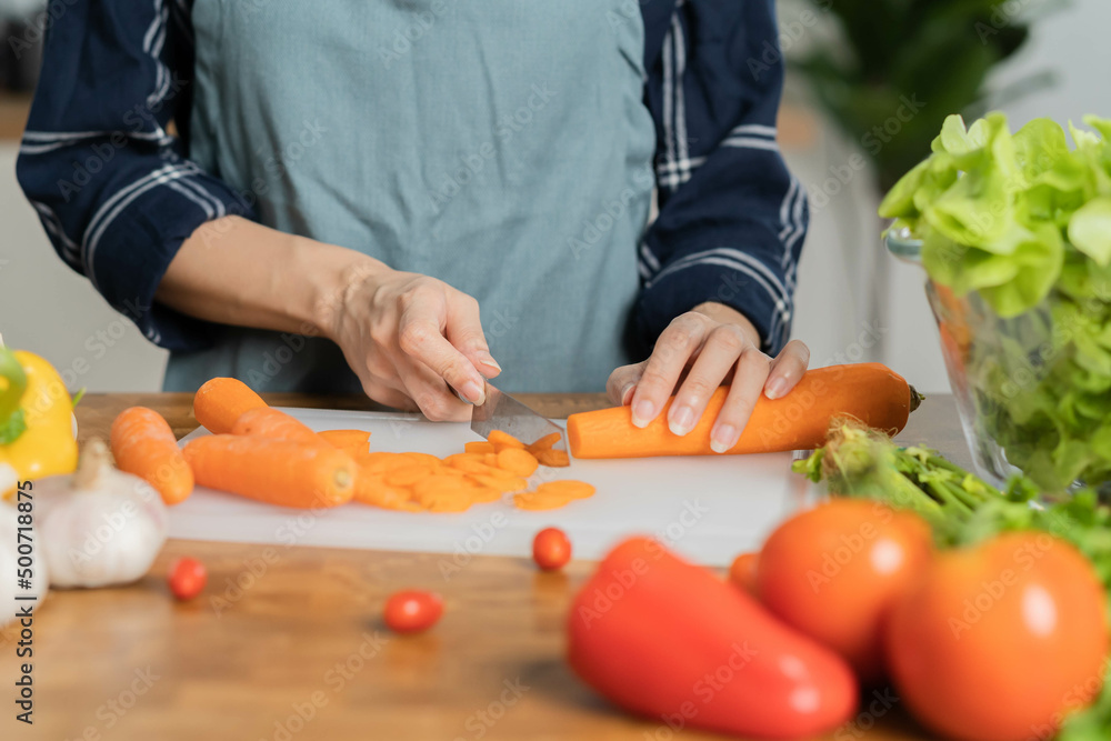 Asian young woman, girl or housewife using knife, cutting carrots on  board, on wooden table in kitchen home, preparing ingredient, recipe fresh vegetables for cooking meal. Healthy food people.