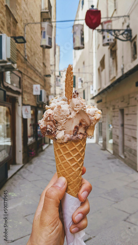 Mano che regge un cono gelato vegano alle creme con cialda biscotto in una via di Alghero, Italia in un giornata di sole photo