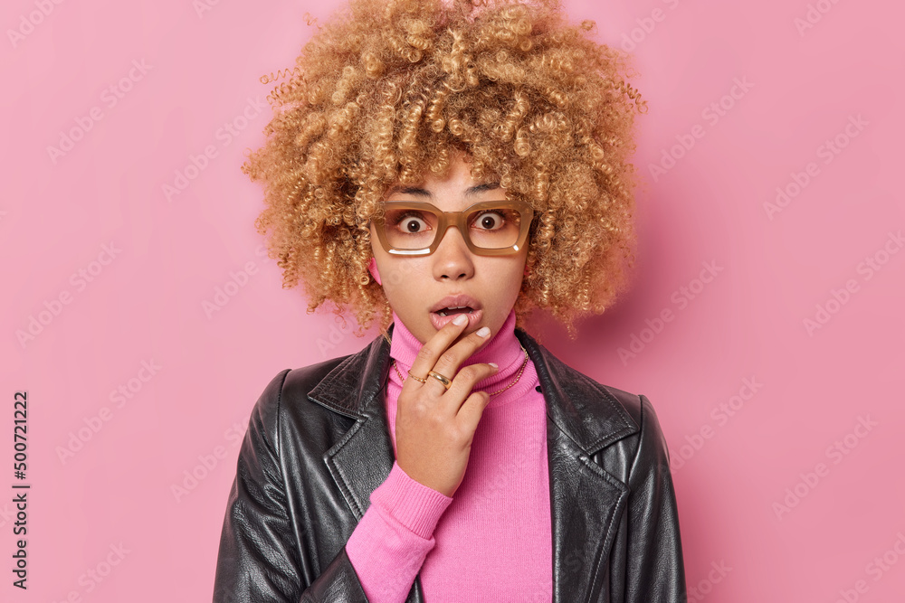 Young beautiful woman feels scared keeps handon chin stares shocked at camera hears amazing news wears spectacles turtleneck and leather jacket isolated over pink background. Negative emotions Young beautiful woman feels scared keeps handon chin stares shocked at camera hears amazing news wears spectacles turtleneck and leather jacket isolated over pink background. Negative emotions