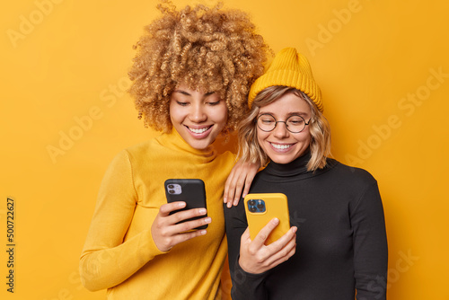Indoor shot of happy beautiful women enjoy mobile offer browse new application or website being addicted to modern technologies holds cellulars chat online isolated over vivid yellow background. photo