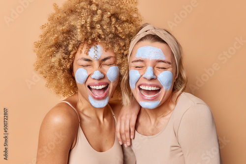 Overjoyed positive young women stand closely to each other keep mouthes opened apply beauty mask for skin care and moisturising dressed casually isolated over brown background. Wellness concept