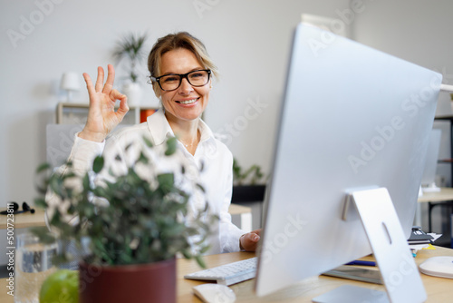 Photography Middle-aged business woman sits at a computer in the office and shows ok gesture