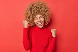 Positive curly haired young woman makes yes gesture celebrates success feels overjoyed and triumphant wears casual turtleneck shakes fists exclaims from joy isolated over vivid red background
