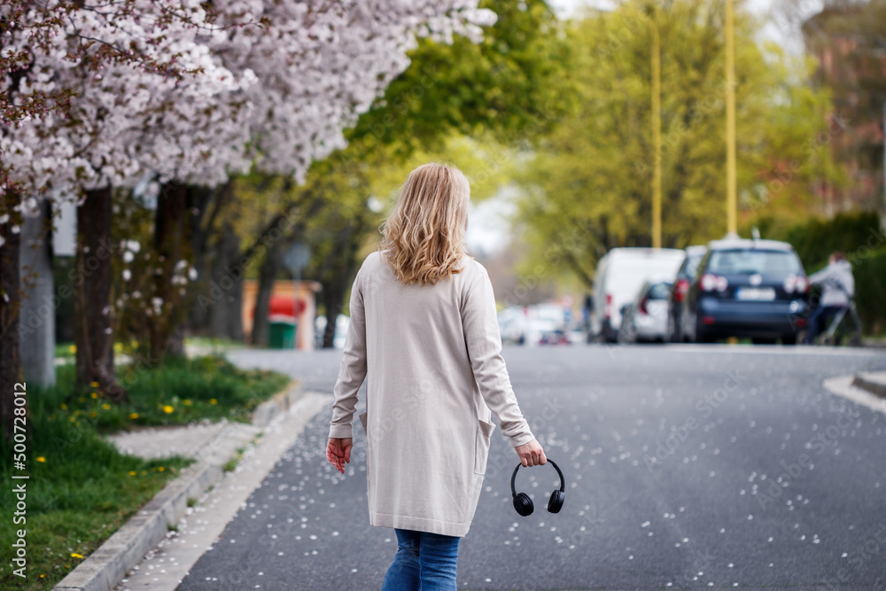 Woman with headphones walking on city street under blooming cherry trees. City life at springtime Woman with headphones walking on city street under blooming cherry trees. City life at springtime