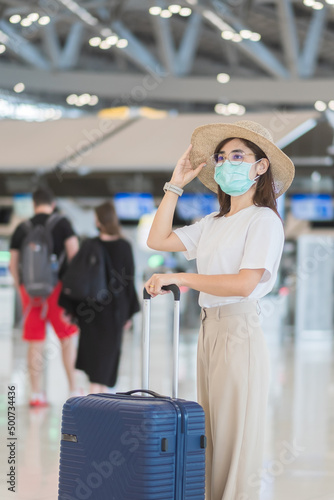 Young female wearing face mask with luggage walking in airport, protection Coronavirus disease infection, Asian woman traveler with hat. Time to travel after vaccine booster dose concept
