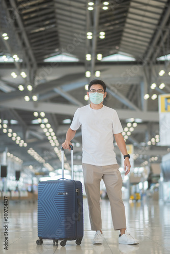 Young male wearing face mask with luggage walking in airport, protection Coronavirus disease infection, Asian man traveler with hat. Time to travel after vaccine booster dose concept