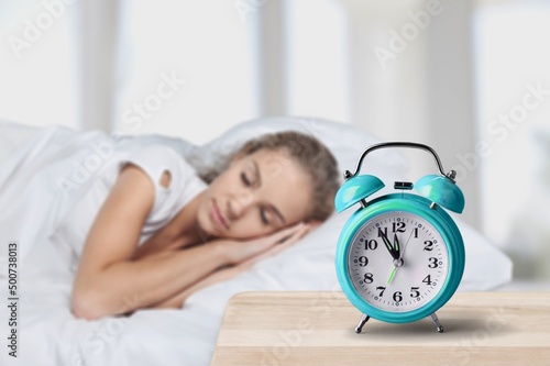 Alarm clock and sleeping young woman in bed. Alarm clock standing on table next to young woman sleeping sweetly in her bed.
