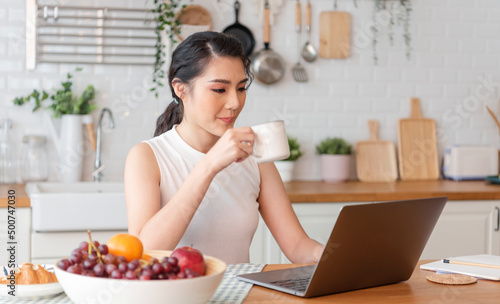 Beautiful young asian woman working on laptop computer while sitting at the kitchen room  drinking coffee.