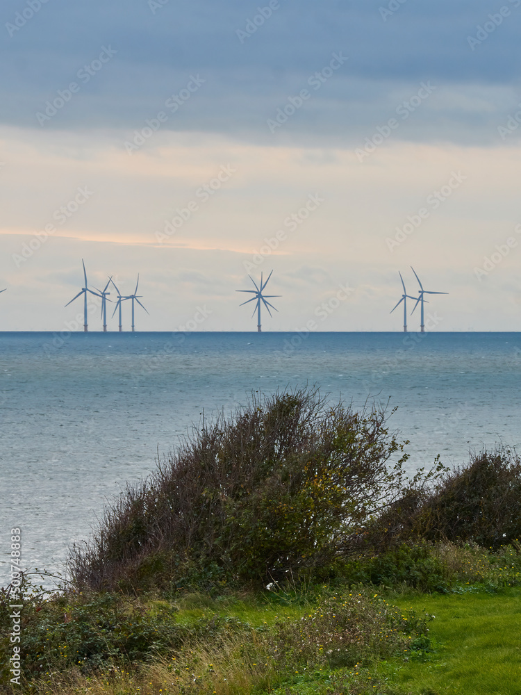 The silhouetted turbines of a wind farm stand on the horizon in the North Sea under a dramatic, stormy sky.