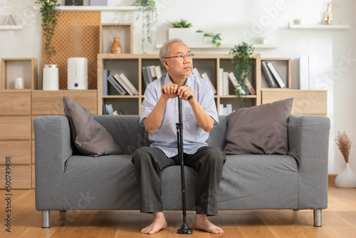 Sad and lonely elderly man sit on couch and looking outside window miss parent and grandchild.Senior asian man holding cane or walking stick sit on sofa alone and sigh deeply stressed at home
