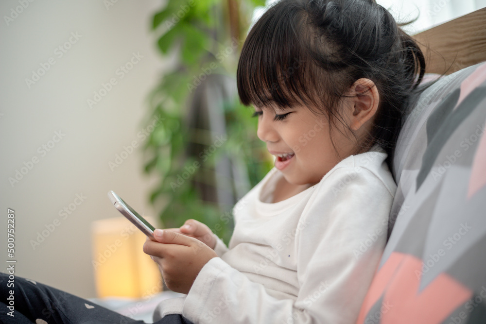 Cute little kid girl watching video on smartphone with smiley face alone on the bed, child using mobile phone with happy face at home. Stay at home quarantine coronavirus COVID-19 pandemic prevention. Cute little kid girl watching video on smartphone with smiley face alone on the bed, child using mobile phone with happy face at home. Stay at home quarantine coronavirus COVID-19 pandemic prevention.
