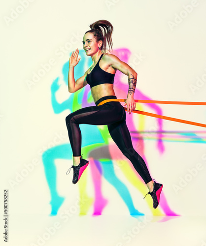 Sporty woman doing jumping exercise with resistance band. Photo of muscular woman in black sportswear on white background with effect of rgb colors shadows.