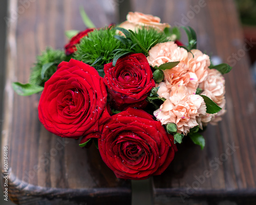 A small bouquet of red roses, delicate pink carnations and green leaves, on a wooden brown background view from above
