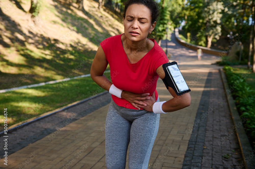 Fototapeta premium Female runner feeling exhaustion and side cramps, stomach-ache during running in the city park. Healthy active lifestyle concept, sport injury