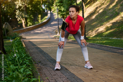 Female runner, Hispanic sportswoman resting in the city park while feeling exhausted during jogging on the treadmill on a warm sunny summer day. Body weight training, slimming and dieting concept