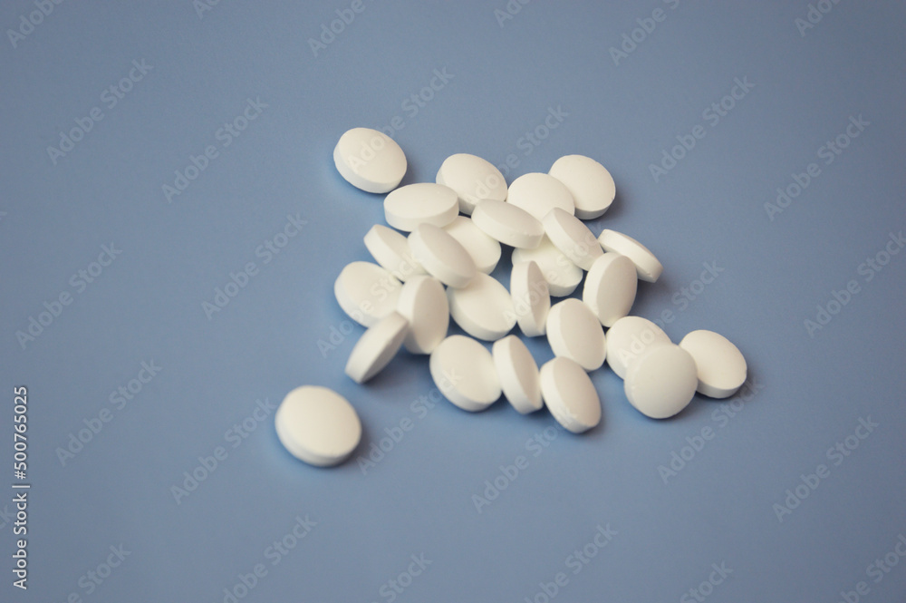 A bunch of simple tiny round white pills scattered on a blue background. Close-up, focus on one pill. The concept of medicine, nutritional supplements and drugs. Free space for text.