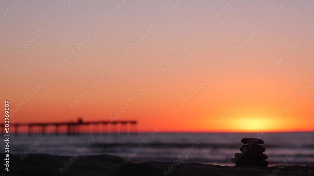 Fototapeta premium Stack of pebble stones, sandy ocean beach, sunset sky. Rock balancing in sun light, sea water waves. Stones staking in pyramid pile. Zen meditation and harmony in balance. Calm tranquil atmosphere.
