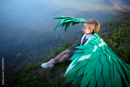 Cute girl in in a grey sweater  white scarf and green wings in nature landscape near wate of lake of river on sunny autumn. Model poses in nature landscape as elf or angel. Unusual photo shoot