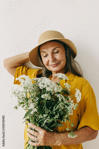 Elderly woman with enjoy blooming bouquet of flowers on beige background. Mature lady in hat, spring vibes. Growing old, beauty of oldness, inspiration, wildflowers concept