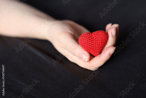 Red knitted heart in palm of female hand on a bed with black bedding. Concept of love, health care, motherhood, blood donation