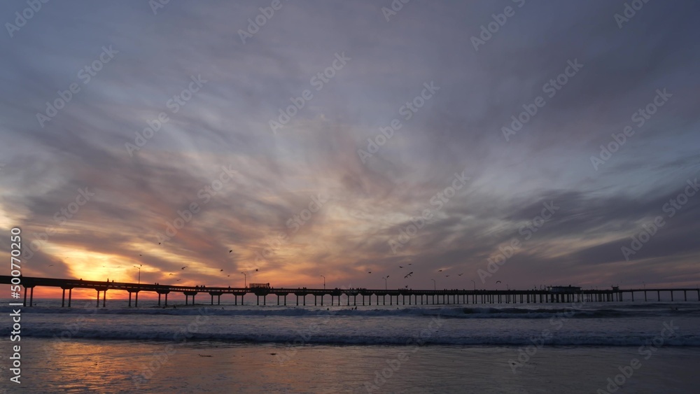 Silhouette of people walking, pier on piles in sea water. Ocean waves, dramatic sky at sunset. California coast aesthetic, beach or shore vibe at sundown. Summer seascape in San Diego near Los Angeles