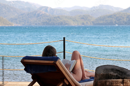 Fototapeta Naklejka Na Ścianę i Meble -  Woman in bikini reading book sitting in deck chair on a sea beach. Summer relax on mountain coast background