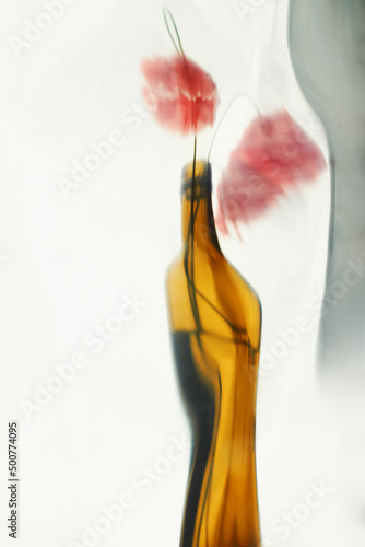 Blurry image of three red poppy flowers in a transparent curved vase on a light background