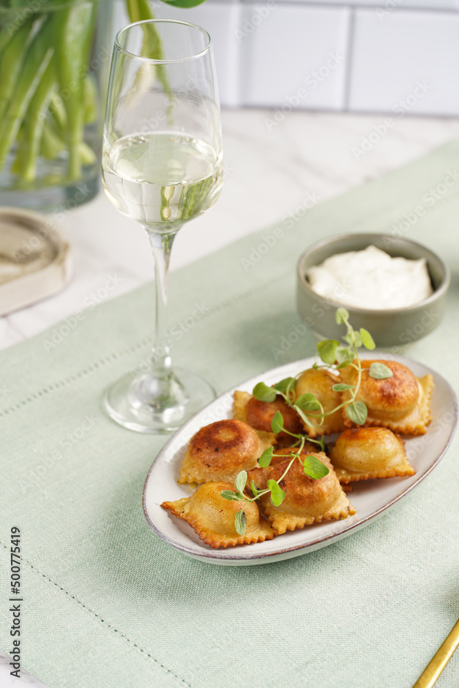 Deep fried traditional italian dish - ravioli, dumplings stuffed with minced meat, oregano branch, sour cream, a glass with champagne on green table cloth