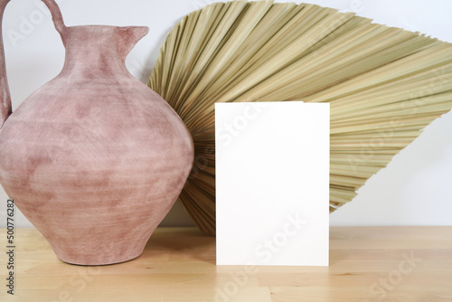 Scandinavian Boho Theme Product Mockup. Empty 5 x 7 greeting card, party invitation with modern beige ceramic vases with dried palm frond leaf against a white wall background.