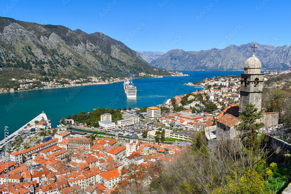 Kotor, Montenegro, Europe. Bay of Kotor on Adriatic Sea. Church, roofs of the historical buildings in the old town. Bay and mountains in the background. Clear blue sky, sunny day