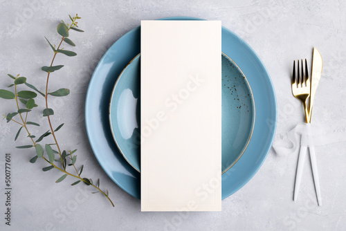 Vertical menu card mockup with festive wedding or birthday table setting with golden cutlery, eucalyptus, blue ceramic plate on grey background. Restaurant menu concept. Flat lay, top view