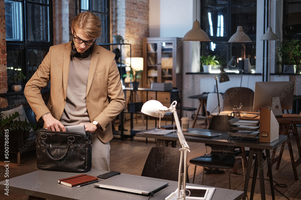 Serious young blond-haired manager with headphones around neck putting tablet into bag while leaving office Serious young blond-haired manager with headphones around neck putting tablet into bag while leaving office