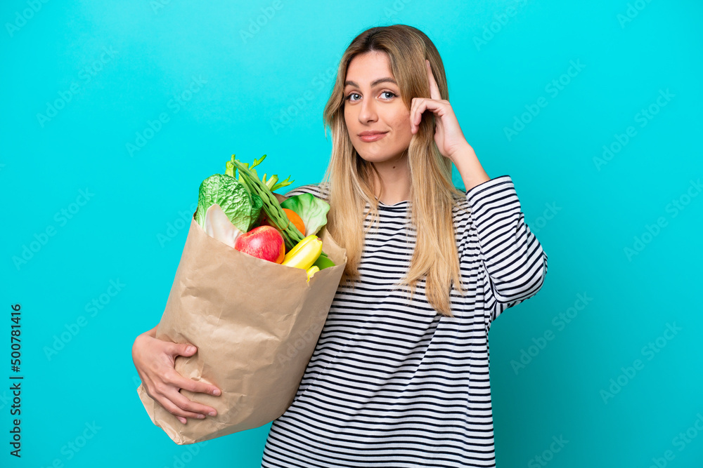 Young Uruguayan woman holding a grocery shopping bag isolated on blue background thinking an idea Young Uruguayan woman holding a grocery shopping bag isolated on blue background thinking an idea