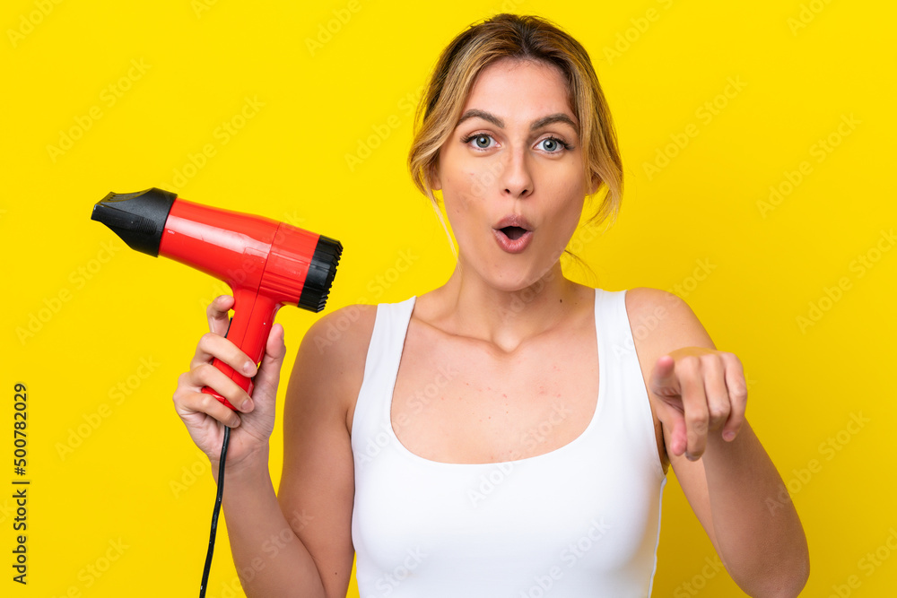 Young Uruguayan woman holding a hairdryer isolate don yellow background surprised and pointing front Young Uruguayan woman holding a hairdryer isolate don yellow background surprised and pointing front