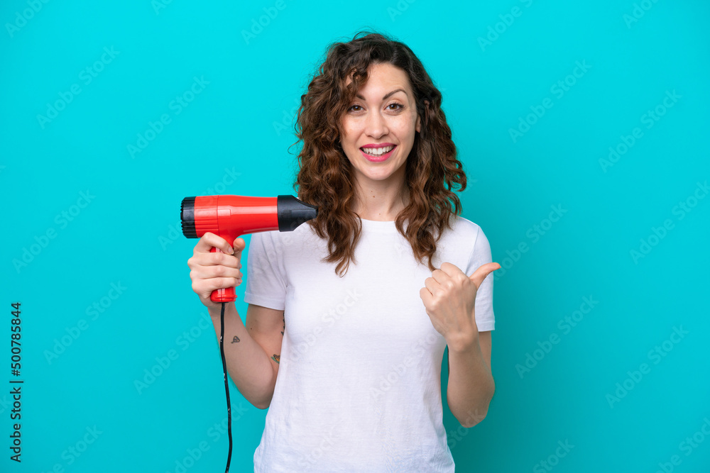 Young caucasian woman holding a hairdryer isolated on blue background pointing to the side to present a product Young caucasian woman holding a hairdryer isolated on blue background pointing to the side to present a product