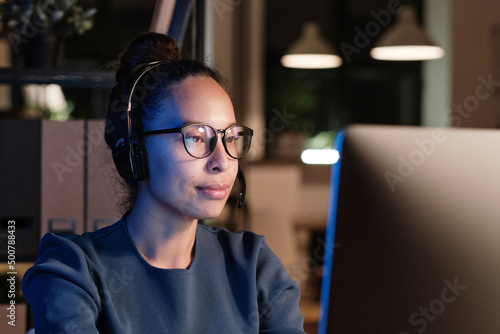 Female customer service specialist in headset with microphone sitting in front of computer in dark office and answering call at night