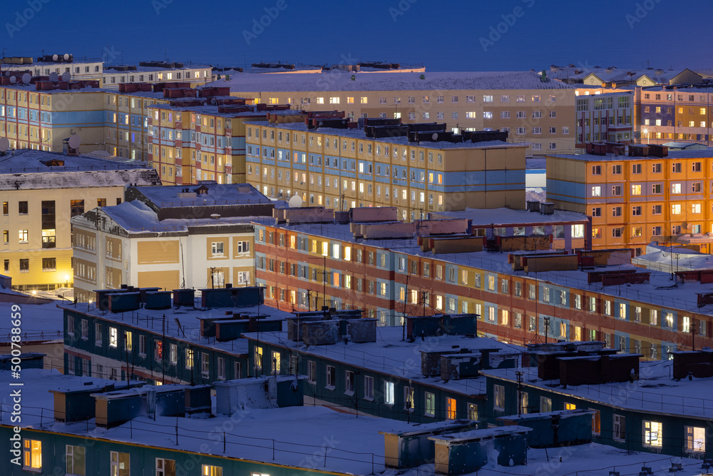 Fototapeta premium Night view from above on a snow-covered northern city in the Arctic. Snow on the roofs of colorful buildings. Cold winter weather. Harsh polar climate. Anadyr, Chukotka, Siberia, Far North of Russia.